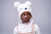 Serious African American little girl in stylish clothes and funny hat standing looking away against gray background — Stock Photo