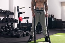 Konzentrierter hemdsloser Sportler, der tagsüber beim funktionellen Training im Fitnessstudio mit Seilen steht — Stockfoto