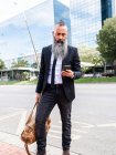 Hombre barbudo de confianza en traje elegante navegando en el teléfono inteligente mientras está de pie cerca de la carretera en la calle con edificios modernos en la ciudad - foto de stock