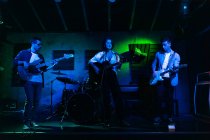 Gruppe von Menschen in lässiger Kleidung, die Gitarre und Schlagzeug spielen, während eine Frau im Club mit Neonlichtern singt und singt — Stockfoto