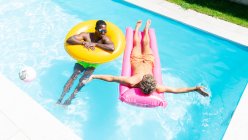 Зверху багаторасові друзі чоловічої статі в басейні, лежачи на надувному кільці і матраці, купаючись в басейні в сонячний літній день — стокове фото