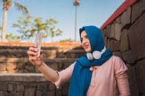 Charmante Muslimin mit traditionellem Kopftuch steht in der Nähe einer Steinmauer und macht an sonnigen Tagen Selbstaufnahmen auf dem Smartphone — Stockfoto