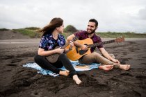 Полная длина улыбающегося мужчины, играющего на акустической гитаре с позитивной подругой, играющей на укулеле, сидя на песчаном побережье в пасмурный день — стоковое фото