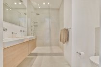 Белые раковины на стене с зеркалом, расположенным рядом со стеклянной душевой кабиной в светлой просторной ванной комнате с инструментами и яркой подсветкой — стоковое фото