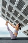 Vista laterale del corpo completo della donna sportiva che pratica Urdhva Mukha Paschimottanasana postura sulla strada vicino al pannello solare moderno durante la sessione di yoga — Foto stock
