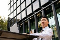 Jeune entrepreneur asiatique poignant avec tasse de boisson chaude et netbook regardant loin à la table de cafétéria urbaine en plein jour — Photo de stock