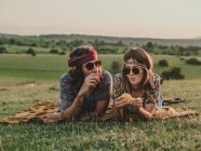 Casal Hippie com óculos de sol descascando e comer uma laranja enquanto deitado no prado gramado na natureza ao pôr do sol — Fotografia de Stock