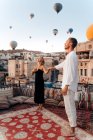 Cuerpo completo de pareja descalza bailando juntos en la terraza de la azotea contra globos de aire caliente volando en cielo despejado - foto de stock
