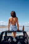 Corpo inteiro de jovem mulher feliz em roupa de verão sentado perto de cerveja no telhado do carro de safári na costa do rio — Fotografia de Stock