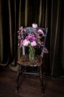 Buquê de peônias coloridas frescas e crisântemos em vaso de vidro colocado em cadeira de madeira desgastada perto de cortinas na sala de luz — Fotografia de Stock