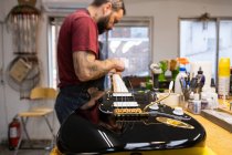 Side view of crop concentrated craftsman with tattoos standing and repairing electric guitar in workshop in daytime — Stock Photo