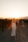Donna bionda in abito bianco guardando lontano con mandria di cavalli in campo sotto il tramonto — Foto stock