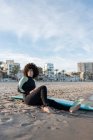 Вид збоку повне тіло босоніж жіночого серфера в гідрокостюмі, що сидить на піщаному пляжі біля моря, що махає — стокове фото