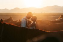 Man embracing tender woman standing close looking at each other among calm horses in hilly countryside in sunset light — Stock Photo