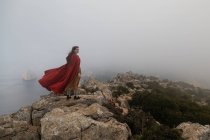 Полный вид на тело женщины в викторианской одежде, стоящей на скале с мохом возле моря в туманную погоду — стоковое фото
