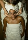 From above of female masseur using massagers while massaging face of client in towel on couch in bright spa center — Stock Photo