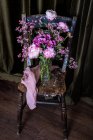 Букет свіжих різнокольорових півоній і хризантем у скляній вазі розміщений на обвітреному дерев'яному стільці біля штор у світлій кімнаті — стокове фото