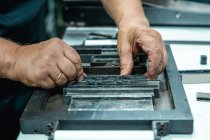 High angle of crop unrecognizable elderly male artisan in apron and eyeglasses preparing metal frame for letterpress printing in studio — Stock Photo