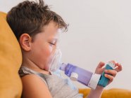 Vue latérale du contenu garçon malade utilisant un respirateur et respirant l'oxygène de l'inhalateur tout en étant assis sur le canapé à la maison — Photo de stock