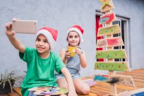 Los niños optimistas que toman autorretrato en el teléfono inteligente mientras están sentados cerca del árbol de Navidad de madera en la habitación durante la preparación de las vacaciones juntos en casa - foto de stock