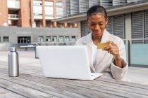 Mujer con tarjeta de crédito en la mano escribiendo en netbook moderno al hacer la compra en línea en la calle en la ciudad - foto de stock