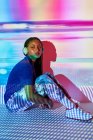 Vista lateral da moda jovem Dominicana millennial feminino com longas tranças afro sentado no chão e olhando para o lado enquanto ouve música em fones de ouvido no quarto com iluminação geométrica colorida — Fotografia de Stock