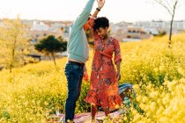 Feliz joven pareja diversa cogida de la mano mientras bailan juntos en el prado floreciente durante un picnic romántico en un día soleado - foto de stock
