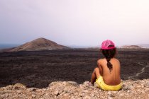 Vue arrière d'un enfant anonyme torse nu admirant la lave volcanique et la mer alors qu'il était assis à Lanzarote Îles Canaries Espagne — Photo de stock