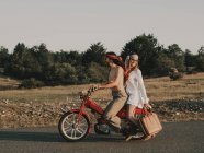 Vista lateral do corpo inteiro do casal hippie montando no ciclomotor vermelho na estrada de asfalto durante a viagem na natureza com árvores no dia de verão — Fotografia de Stock