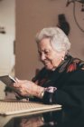 Mujer anciana sonriente usando ropa de abrigo sentada en la mesa con la tableta y la taza de té mirando a la pantalla - foto de stock