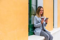 Красивая женщина в стильном наряде с кофеваркой смотрит в камеру, пока текст на мобильном телефоне рядом со зданием с кошельком — стоковое фото