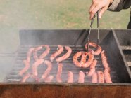 Cropped unrecognizable chef with tongs cooking various types of tasty sausages roasting on grill grate above charcoal in countryside during barbecue in countryside on summer day — Stock Photo
