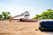 Side view of calm female in white activewear performing Halasana on mat on ground in park against green trees and cloudless blue sky in sunny day — Stock Photo