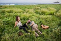 Felices amigos músicos con guitarra y ukelele sentado sobre hierba verde en la costa cerca del océano en la naturaleza durante el día - foto de stock