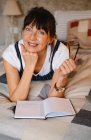 Positive middle age female leaning on hand while lying on bed with opened notepad and eyeglasses and looking at camera — Stock Photo