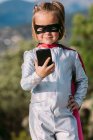 Самоуверенная девушка в костюме супергероя в маске для глаз с накидкой, просматривающей мобильный телефон — стоковое фото