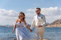 Casal de casamento alegre andando na costa perto do mar ondulante enquanto desfruta do dia do casamento na natureza ensolarada — Fotografia de Stock