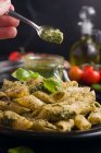 Chef méconnaissable mettant de la sauce au pesto vert sur une assiette avec des pâtes et des feuilles de basilic servies sur une table sur fond noir — Photo de stock