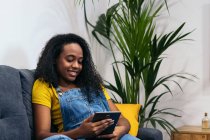 Positive African American woman in denim outfit sitting on couch and smiling while browsing tablet at home — Stock Photo