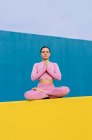 Мирна жінка в рожевому активаторі сидить в Падмасані з руками Намасте і медитує під час сеансу йоги на синьому і жовтому фоні. — стокове фото