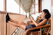 Вид збоку весела молода жінка-туристка в повсякденному одязі посміхається, сидячи за допомогою смартфона на кріслі на дерев'яній терасі старого будинку в сонячний день — стокове фото