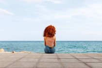 Vista posteriore di femmina irriconoscibile con riccioli di zenzero seduti su argine di pietra e ammirando il paesaggio marino — Foto stock