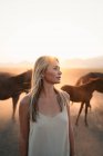 Blonde Frau im weißen Kleid mit Pferdeherde im Feld bei Sonnenuntergang — Stockfoto