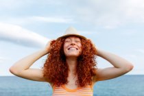 Happy young female with closed eyes putting hat on curly hair on seashore during vacation — Stock Photo