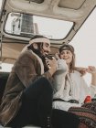Позитивная пара хиппи в костюмах и повязках в стиле бохо, сидящая в старом автомобиле во время поездки на природу в летний день — стоковое фото