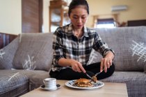 Joven hembra asiática comiendo panqueques caseros colocados en el plato cerca de la taza de café en la mesa mientras está sentado en un cómodo sofá en la sala de estar - foto de stock