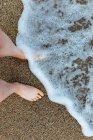 From above of crop unrecognizable barefoot female traveler standing on wet sandy shore washed by foamy wave — Stock Photo