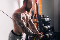 Vista lateral del deportista muscular concentrado con tatuajes haciendo ejercicios en la máquina de cruce de cables en el gimnasio con paredes ligeras - foto de stock