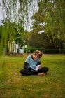 Повне тіло концентрованої босоніжки читає цікаву книгу, сидячи на трав'янистому газоні на подвір'ї проти житлового будинку в сільській місцевості — стокове фото