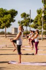 Вид сбоку на группу женщин в активах, стоящих коленом к груди на циновках для йоги во время сессии в парке с зелеными деревьями в солнечный день — стоковое фото